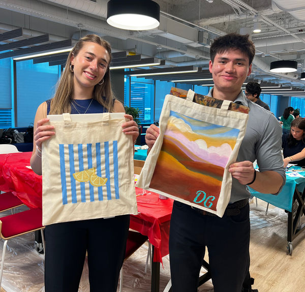 Two colleagues show the tote bags they painted, one with blue stripes and lemons, one with a sunset