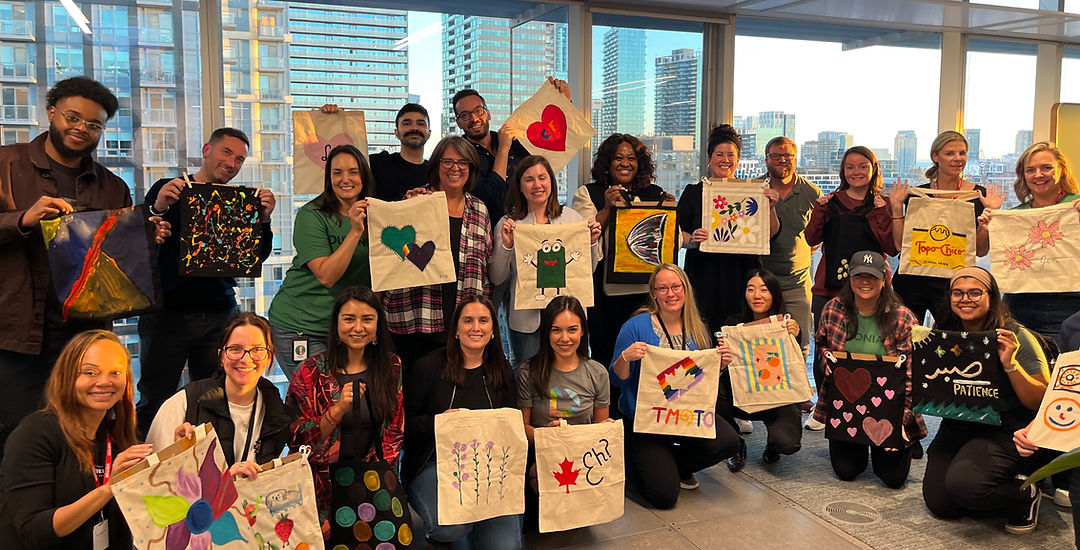 An office team holds up the tote bags they painted together, with city towers behind them