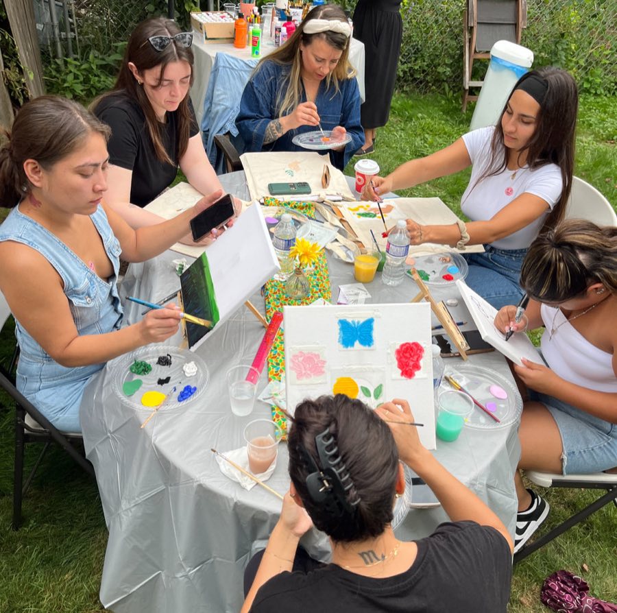 Friends around a table in a backyard, painting canvases and tote bags