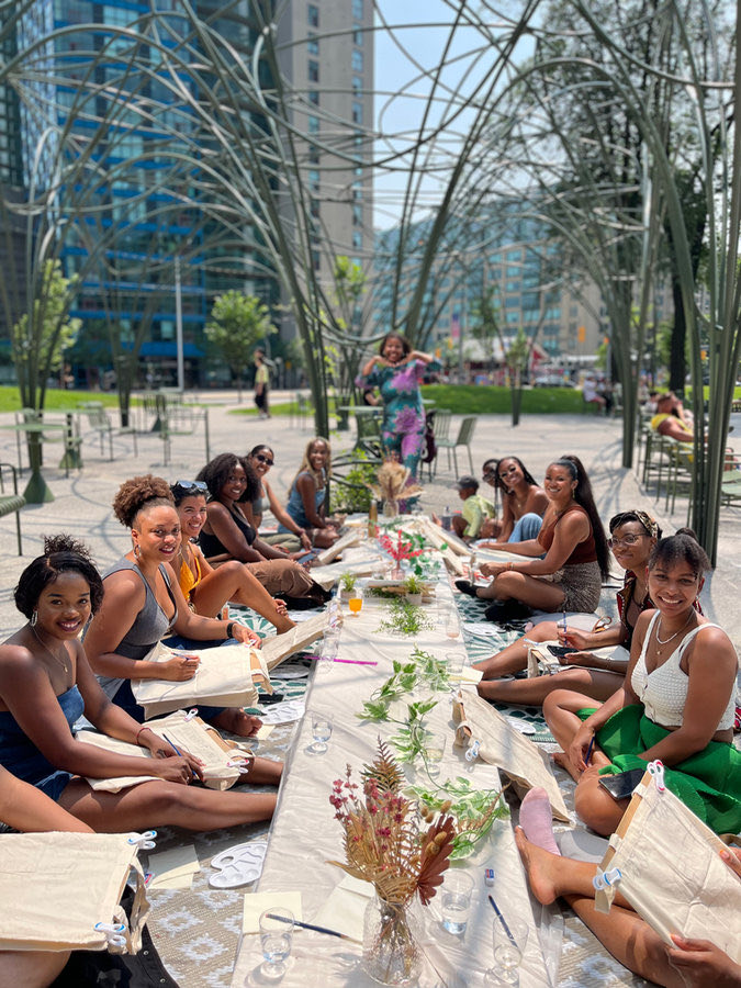 Guests sit along a long table set with flowers and greenery outdoors, each with a tote bag to paint