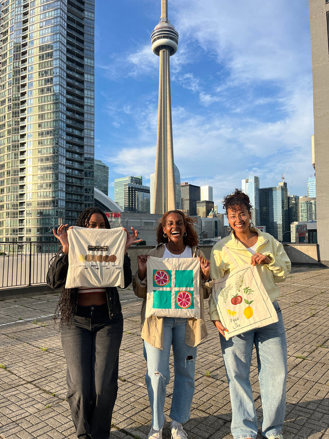Three friends hold up their painted tote bags in front of the CN Tower